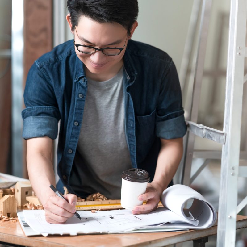 asian attractive glasses interior designer working at house site renovation construction progress with smile and confident at desk with blue print and drawing tool